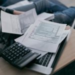 A young adult using a calculator and laptop to manage finances at home, surrounded by papers.