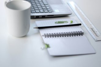 black pencil on ruled notepad beside white ceramic mug and gray laptop computer